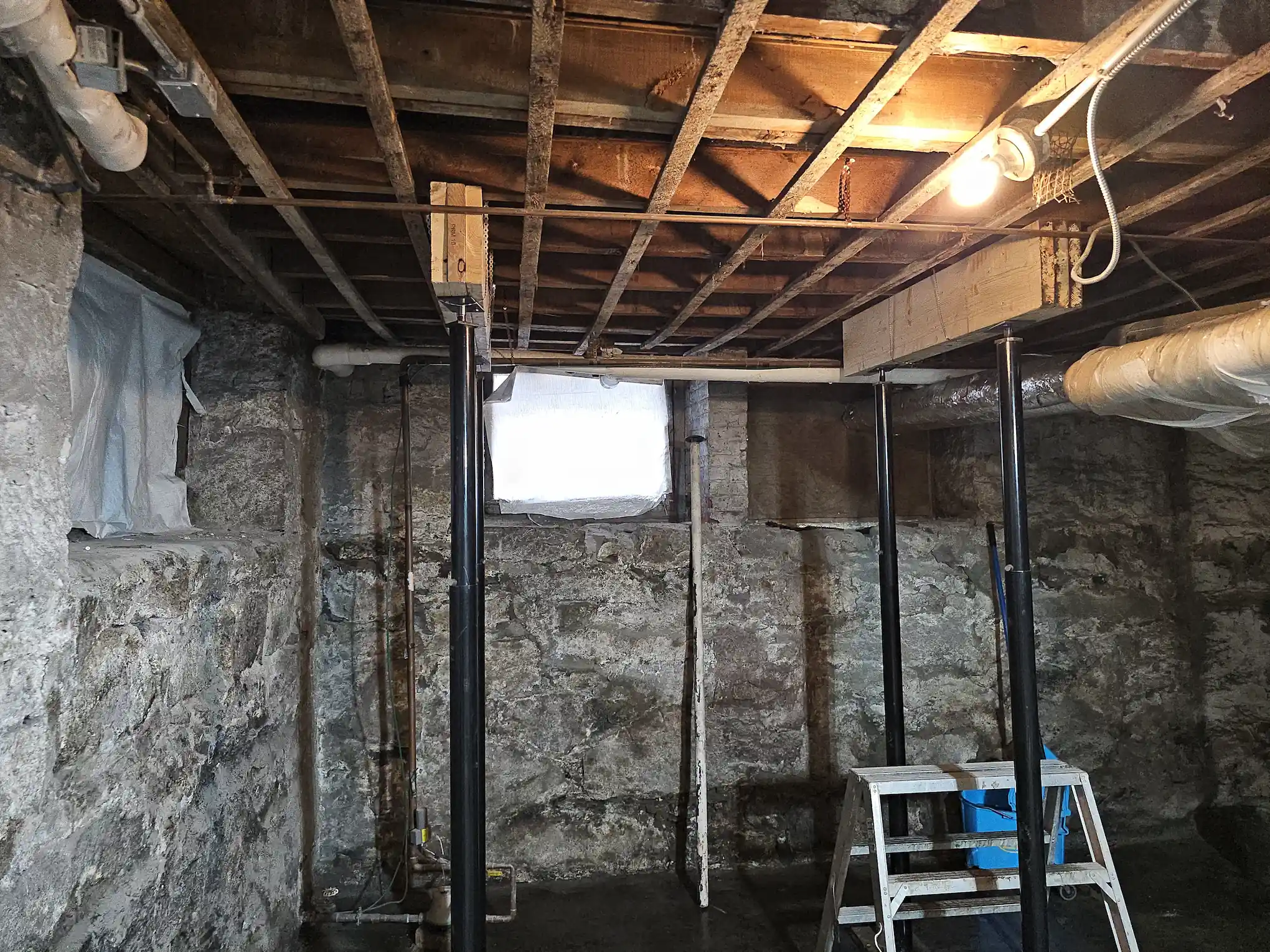 A basement under renovation with exposed stone walls, wooden beams, support columns, and a ladder.