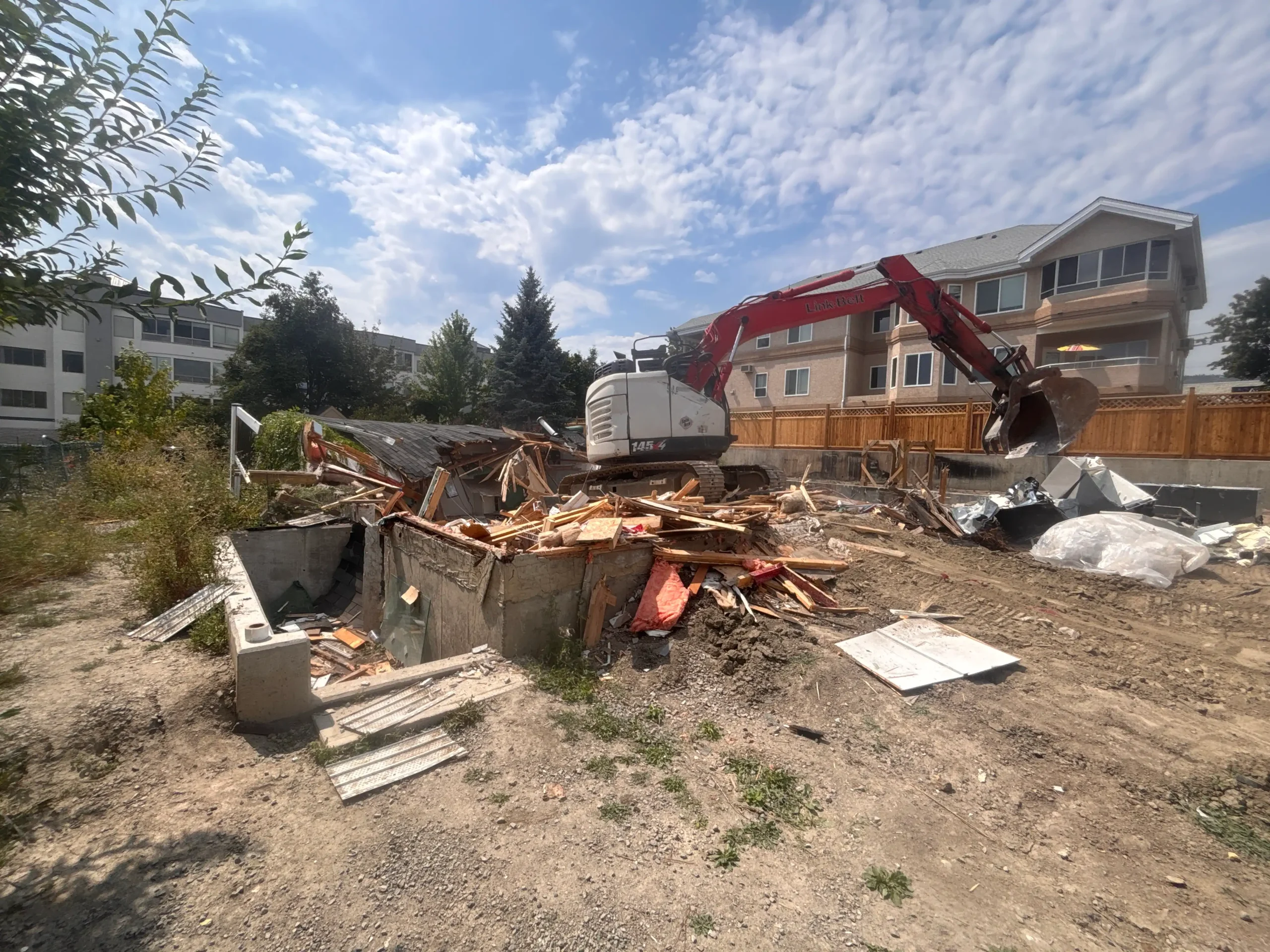 A demolition site with an excavator removing debris, surrounded by scattered wood and materials under a partly cloudy sky.