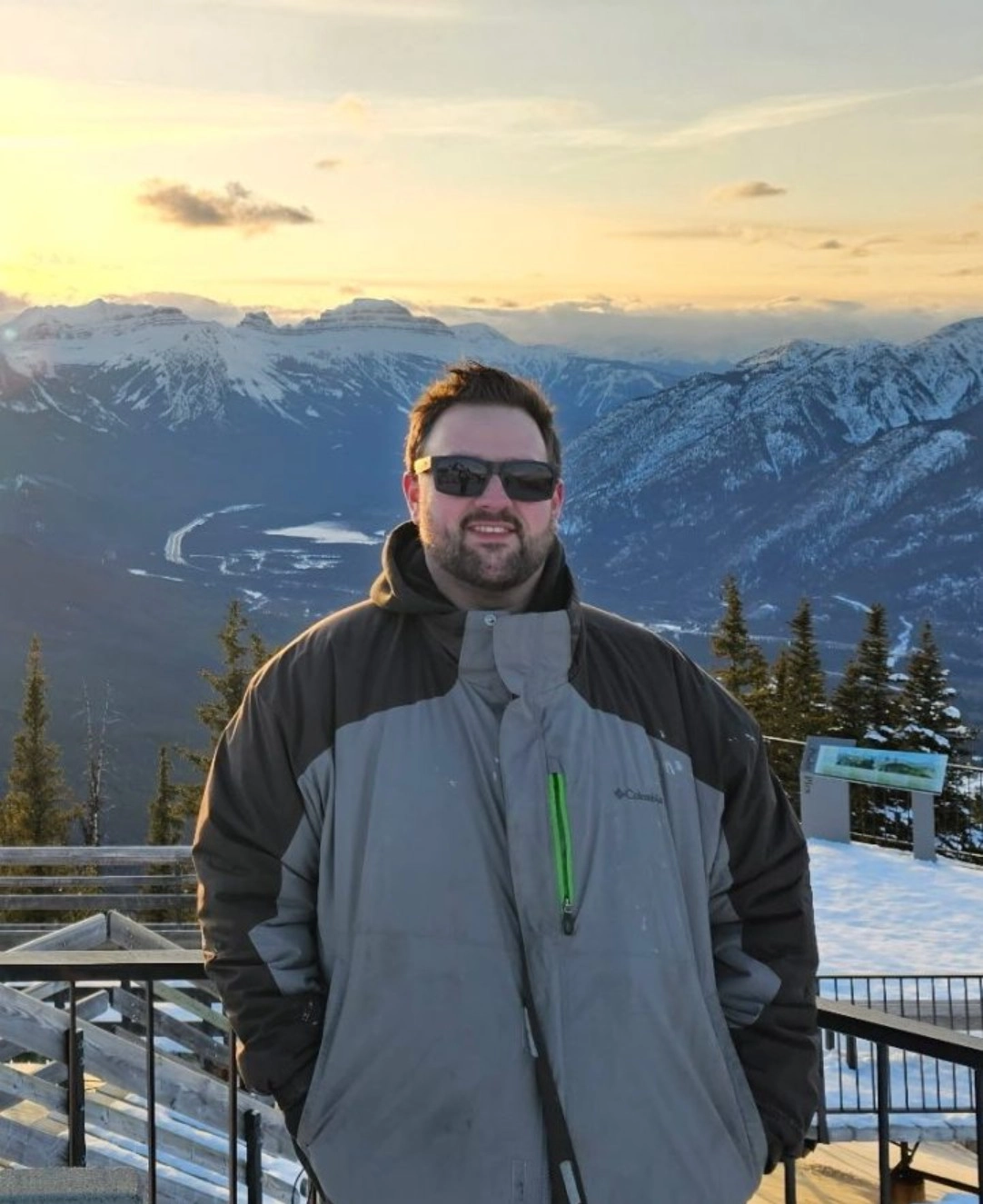 A man in a winter jacket standing at a mountain viewpoint with snow-covered peaks and a sunset sky in the background.