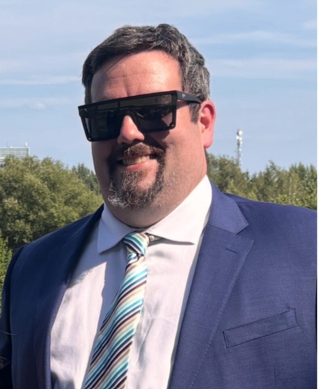 A man wearing a blue suit, white shirt, and striped tie, standing outdoors with trees and a clear sky in the background.